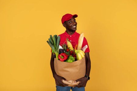 Photo for Curious smiling millennial black guy courier hold paper bag with products, look at free space, isolated on orange studio background. Food delivery at home, work and service - Royalty Free Image