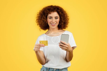 Photo for Smiling teen caucasian curly lady with credit card pays with smartphone for order, isolated on yellow studio background. Shopaholic lifestyle, sale, online shopping and banking, finance - Royalty Free Image
