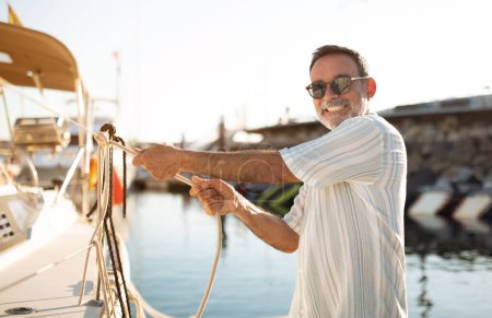 Foto de Harbor Skills. El hombre mayor gestiona con confianza la cuerda, atracando su yate en el puerto deportivo, sonriendo a la cámara mientras demuestra años de experiencia en navegación en el muelle iluminado por el sol. Ocio de vacaciones junto al mar - Imagen libre de derechos