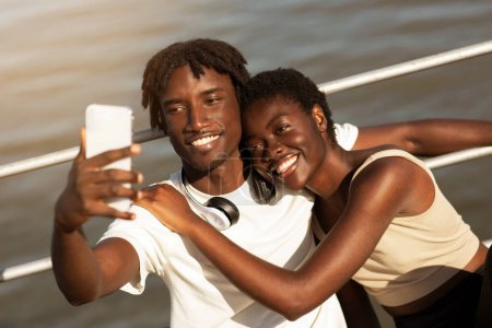 Photo for Happy Black Millennial Couple Taking Selfie On Smartphone While Doing Sport Together Outdoors, Smiling Young African American Man And Woman Looking At Device Camera And Embracing, Closeup Shot - Royalty Free Image