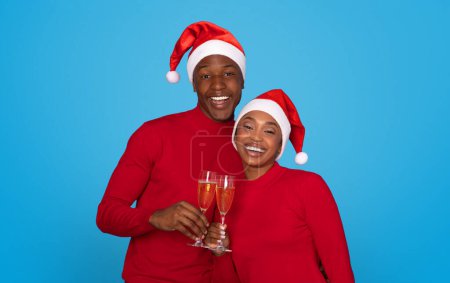 Photo for Joyful Black Couple In Santa Hats Toasting With Champagne Glasses Against Blue Background In Studio, Celebrating Xmas Holiday Season And Exchanging Warm Festive Wishes, Smiling To Camera - Royalty Free Image