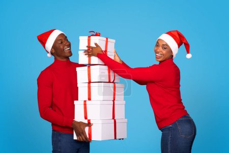 Téléchargez les photos : Jeune couple afro-américain manipulant des piles de boîtes-cadeaux avec des rubans rouges, fond bleu. Conjoints appréciant l'excitation des échanges présents de Noël et des célébrations festives - en image libre de droit