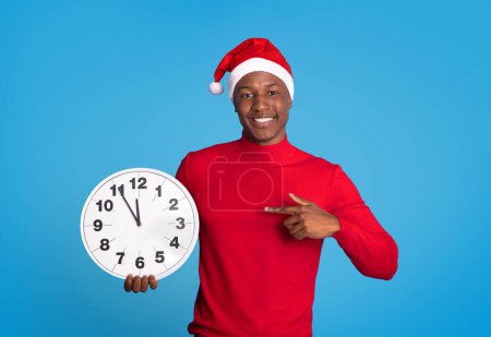 Foto de Hombre negro con sombrero de Santa festivo señalando con el dedo al reloj de pie contra el telón de fondo azul en el estudio, cuenta atrás minutos para el próximo año. Emoción de las festividades de Navidad y Año Nuevo - Imagen libre de derechos