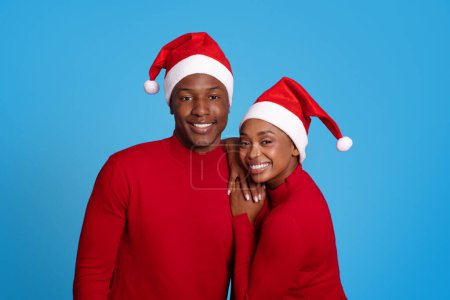 Photo for Black woman and man sharing a close hug over blue studio background, highlighting loving relationship during the holiday season, wearing red Santa hats. New Year Celebration And Winter Season - Royalty Free Image