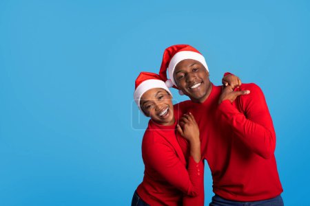 Foto de Alegre esposo y esposa afroamericanos en sombreros de Papá Noel Abrazándose cariñosamente en el fondo azul en el estudio, enfatizando la alegría de estar juntos durante las vacaciones de Navidad. Copiar espacio - Imagen libre de derechos