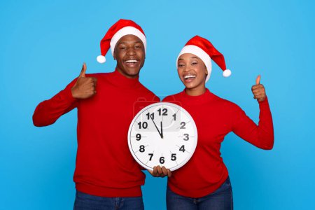 Téléchargez les photos : Joyeux jeunes conjoints noirs dans Santa Hats tenant horloge sur fond de studio bleu, Gesturing Thumbs Up approuvant le Nouvel An à venir. Nous aimons Noël vacances Time Concept - en image libre de droit