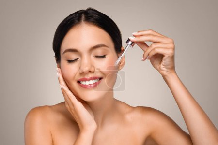 Photo for Cheerful young caucasian naked woman apply serum to skin face, enjoy spa treatments at home, isolated on gray studio background. Beauty care, anti-wrinkle, anti aging procedures - Royalty Free Image