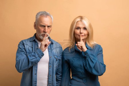 Téléchargez les photos : Mystérieux homme et femme à la retraite montrant un geste silencieux garder le doigt sur les lèvres et regarder la caméra, isolé sur fond de studio beige. Couple sénior garder le secret - en image libre de droit