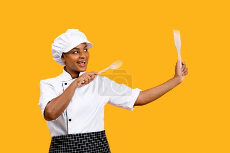 Téléchargez les photos : Jeune chef noir ludique dame avec spatules dans les mains posant sur fond jaune studio, Happy African American Cook femme portant uniforme s'amuser avec des ustencils de cuisine, Copier l'espace - en image libre de droit