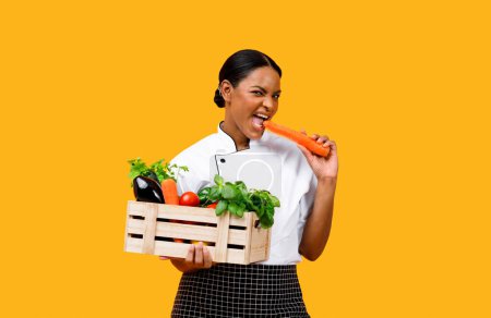 Foto de Chef negro mujer sosteniendo jaula de madera con verduras orgánicas y zanahoria mordida, alegre señora cocinera afroamericana disfrutando de sabrosos ingredientes frescos para cocinar, de pie sobre fondo amarillo - Imagen libre de derechos