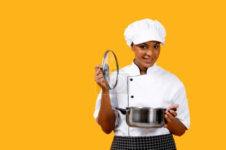 Photo for Nice Recipe. Happy Black Chef Woman Opening Saucepan And Smiling At Camera, Beautiful Young African American Cook Lady In Uniform Standing Isolated Over Yellow Studio Background, Copy Space - Royalty Free Image