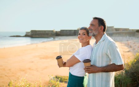 Téléchargez les photos : Des moments de vacances. Homme mûr enroulant ses bras autour de sa femme debout près de l'océan, les deux tenant des tasses à café en papier. Couple sénior romantique ayant des vacances d'été parfaites par le littoral. Espace pour le texte - en image libre de droit