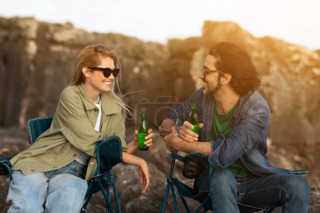 Téléchargez les photos : Joyeux jeune couple buvant des boissons gazeuses tout en se relaxant ensemble à l'extérieur, joyeux homme et femme millénaire bavardant et riant, se reposant dans des chaises de camping sur des rochers de plage, gros plan - en image libre de droit