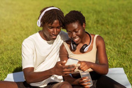 Photo for Young Black Sporty Couple Using Fitness App On Smartphone After Training Outdoors, Happy African American Man And Woman Browsing Sport Tracker Application While Relaxing After Outside Workout - Royalty Free Image