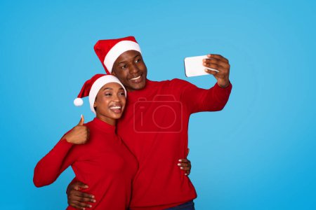 Téléchargez les photos : Des conjoints noirs souriants dans des chapeaux de Père Noël posant pour selfie à l'aide d'un smartphone, étreignant et gestualisant les pouces sur fond de studio bleu. Homme et femme prenant des photos festives et salutations de Noël - en image libre de droit