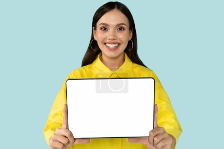 Téléchargez les photos : Joyeux joyeux joyeux jeune femme brune portant chemise jaune montrant tablette numérique avec écran blanc vide et souriant à la caméra, recommandant belle application mobile, offre en ligne, maquette - en image libre de droit