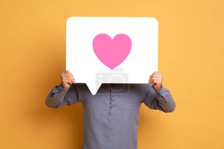 Foto de Hombre con camisa gris oculta su cara mientras presenta una gran burbuja del habla que contiene un corazón rosado vibrante sobre un fondo amarillo, que simboliza el amor o la apreciación - Imagen libre de derechos