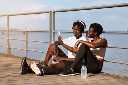 Photo for Fitness App. Motivated Young Black Couple Checking Their Sport Activity On Smartphone After Training Outdoors, African American Man And Woman Sitting On Pier Near Sea, Browsing Mobile Application - Royalty Free Image