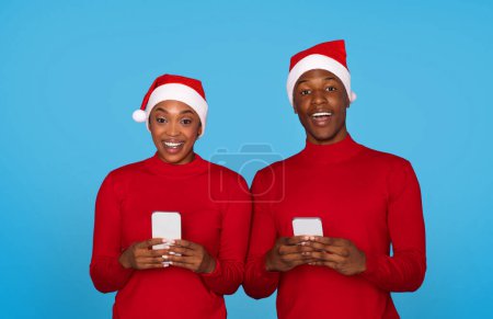 Téléchargez les photos : Couple noir joyeux chapeaux Santa en utilisant les téléphones cellulaires et souriant à la caméra sur fond bleu studio. Homme et femme textos et navigation sur Internet sur les smartphones modernes. Offre mobile pour Noël - en image libre de droit