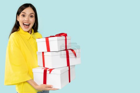 Photo for Excited pretty brunette lady in yellow shirt holding stack of gift boxes over blue studio background. Celebrating Womans Day, Valentines, birthday, anniversary. Shopping for holiday, copy space - Royalty Free Image