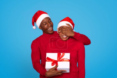 Photo for Christmas Gift. African American Man In Santa Hat Surprising Wife, Presenting Romantic Xmas Gift And Covering Womans Eyes Over Blue Background, Studio Shot. Winter Holiday Advertisement - Royalty Free Image