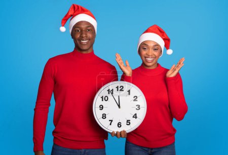 Photo for African American Couple Holding Large Clock On Bright Blue Background In Studio, Signifying Countdown To Upcoming New Year And Embracing Joy Of Christmas Celebration Together - Royalty Free Image