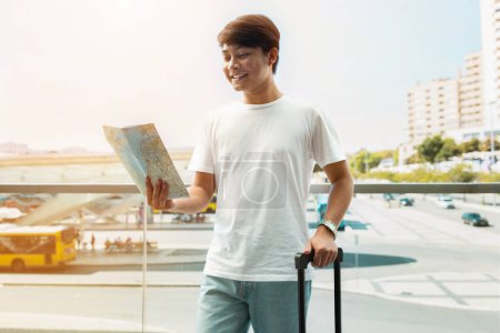 Téléchargez les photos : Heureux jeune homme asiatique touriste est arrivé à destination. Poser à l'extérieur à côté de l'aéroport avec des bagages, regarder la carte sur papier et sourire, profiter de ses vacances - en image libre de droit
