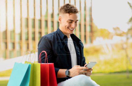 Photo for Happy millennial caucasian man in casual with many bags, chatting by phone, use banking near city mall building, outdoor. Man enjoy online shopping, sale app on gadget, spare time - Royalty Free Image