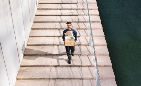 Photo for Arabic business guy holding carton box of belongings walking up steps outside of corporate building, capturing moment of quitting and seeking new career horizons. Full length - Royalty Free Image