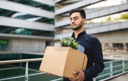 Photo for Dismissal issue. Unhappy fired middle eastern manager guy exiting modern corporate building with personal items in cardboard box. Business layoffs and financial strain. - Royalty Free Image