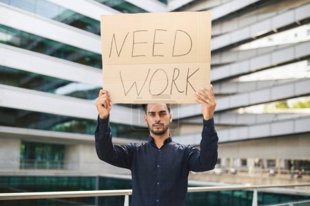 Photo for Unhappy middle eastern businessman holding Need Work paper poster, stands outside of modern office building. Unemployment issue, economic challenges and layoffs concept - Royalty Free Image