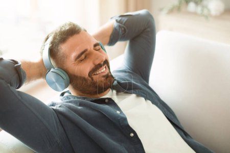 Photo for Relaxation and rest. Relaxed bearded european man closing eyes enjoying music in modern wireless headphones, lying holding hands behind head, resting on sofa at home interior - Royalty Free Image