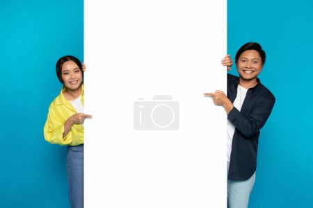 Photo for Positive millennial asian couple in casual point finger at big banner with free space for words, text, ad and offer, isolated on blue studio background. Good news, attention, recommendation sale - Royalty Free Image