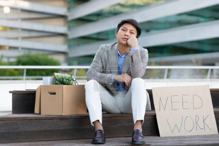 Photo for Unemployment issue. Unhappy fired asian business worker man sitting with paper sign text Need Work and carton box of his belongings, outdoor in urban area. Jobless guy concept - Royalty Free Image