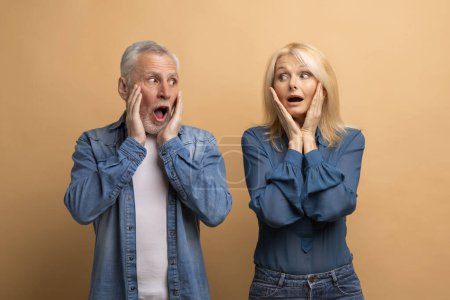 Photo for Shocked emotional senior man and woman looking at each other with aghast face expression, touching their faces and exclaiming, isolated on beige studio background. Shocking news - Royalty Free Image