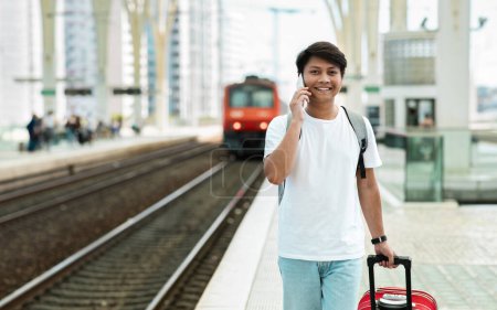 Photo for Cheerful young asian man tourist walking by train station, carrying luggage, talking on phone with friend, calling taxi, arrived to new city, chinese guy traveling, copy space - Royalty Free Image