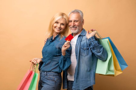 Photo for Happy retired couple shopaholics with colorful shopping bags and credit card posing on beige background. Cheerful senior man and woman enjoying sale season, black friday deal, showing purchases - Royalty Free Image
