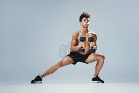 Photo for Athletic young man performing deep lunge while holding dumbbells, exercising on gray studio backdrop, full length. Strength and discipline, gym workout with weights concept - Royalty Free Image