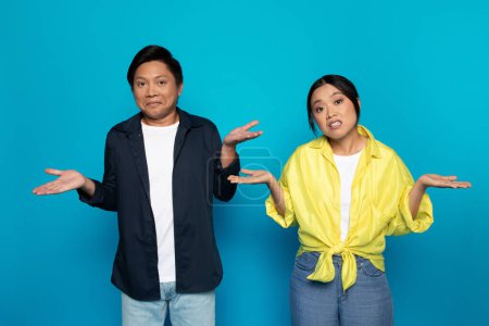 Photo for Sad confused millennial asian couple in casual spreads hands to sides, hold free space, isolated on blue studio background. Emotions, question, choose gesture, feeling doubt - Royalty Free Image