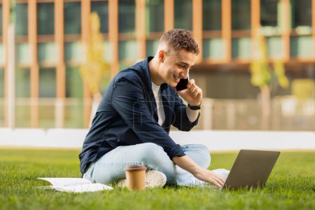 Photo for Smiling busy young european guy in casual enjoy lifestyle, work on laptop, call by phone in park on grass, near office building. Work, business, study with gadget, freelance - Royalty Free Image