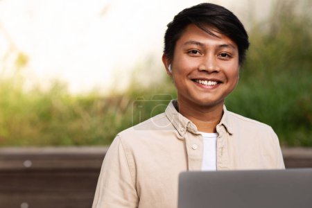 Téléchargez les photos : Positif attrayant asiatique jeune homme portant tenue décontractée assis sur le banc au parc, travailler en ligne, à l'aide d'un ordinateur portable, profiter de la journée ensoleillée, regarder la caméra et sourire, copier l'espace - en image libre de droit