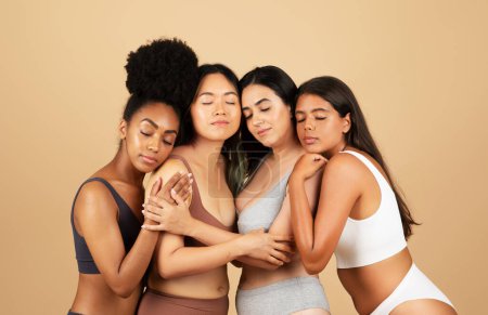 Foto de Cuatro mujeres diversas irradian belleza natural mientras están de pie cerca, vistiendo ropa interior simple y compartiendo un abrazo reconfortante contra un fondo beige neutro - Imagen libre de derechos