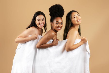 Téléchargez les photos : Trois femmes heureuses et diverses se tiennent côte à côte, confortablement enveloppées dans une serviette blanche, rayonnantes de véritables sourires sur fond beige neutre - en image libre de droit