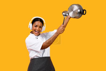 Téléchargez les photos : Happy Young Black Chef Femme dans les écouteurs s'amuser tout en cuisinant, gai afro-américaine Cook Lady utilisant la casserole comme tambour, faire de la musique avec des ustensiles de cuisine, debout sur fond jaune - en image libre de droit