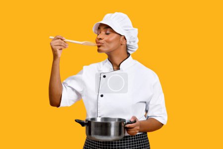 Photo for Happy young black chef woman holding saucepan and wooden spoon, african american cook lady tasting food while cooking, standing isolated over yellow studio background, enjoying preparing meal - Royalty Free Image