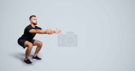 Photo for Focused calm handsome strong millennial european guy with beard in sportswear squat, isolated on gray studio background, full length, panorama. Body care, sport and workout - Royalty Free Image