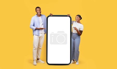 Téléchargez les photos : Cool offre mobile. Heureux homme et femme noirs pointant vers un énorme téléphone portable avec écran blanc, démontrant l'espace libre pour la conception de l'application téléphonique ou du site Web, fond jaune - en image libre de droit