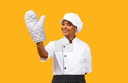 Photo for Happy Young Black Chef Woman Demonstrating At Oven Mitt On Her Hand, Professional African American Cook Lady In Uniform And Hat, Posing Isolated Over Yellow Background, Enjoying Cooking, Copy Space - Royalty Free Image