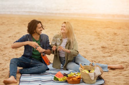 Foto de Feliz joven pareja disfrutando de un picnic en la playa cerca del mar, hombre abriendo una botella de vino mientras su novia sostiene vasos, hombre y mujer europeos románticos teniendo citas al aire libre, espacio de copia - Imagen libre de derechos