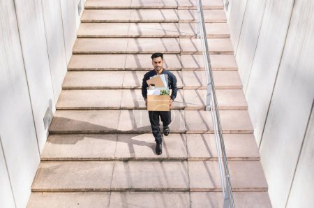 Photo for Young middle eastern businessman carrying cardboard box, walking up modern urban staircase outdoor, symbolizing fresh start in new employment opportunity. Full length, empty space - Royalty Free Image